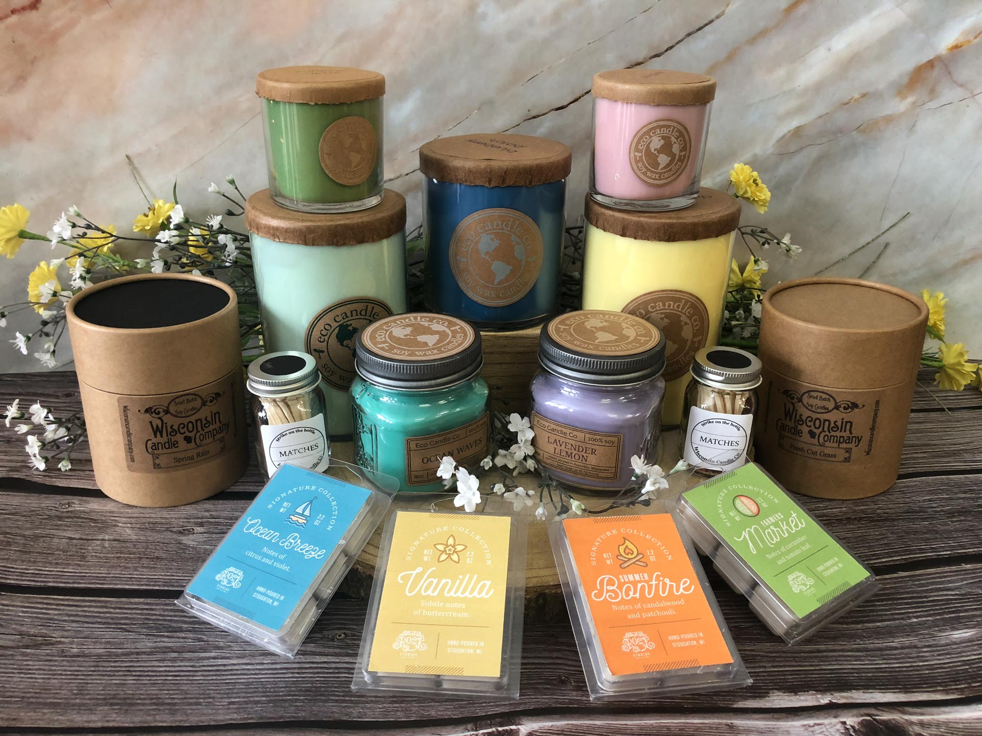 Assorted colorful candles in glass jars and brown containers displayed on a wooden surface with wax melts in front and decorative flowers in the background.