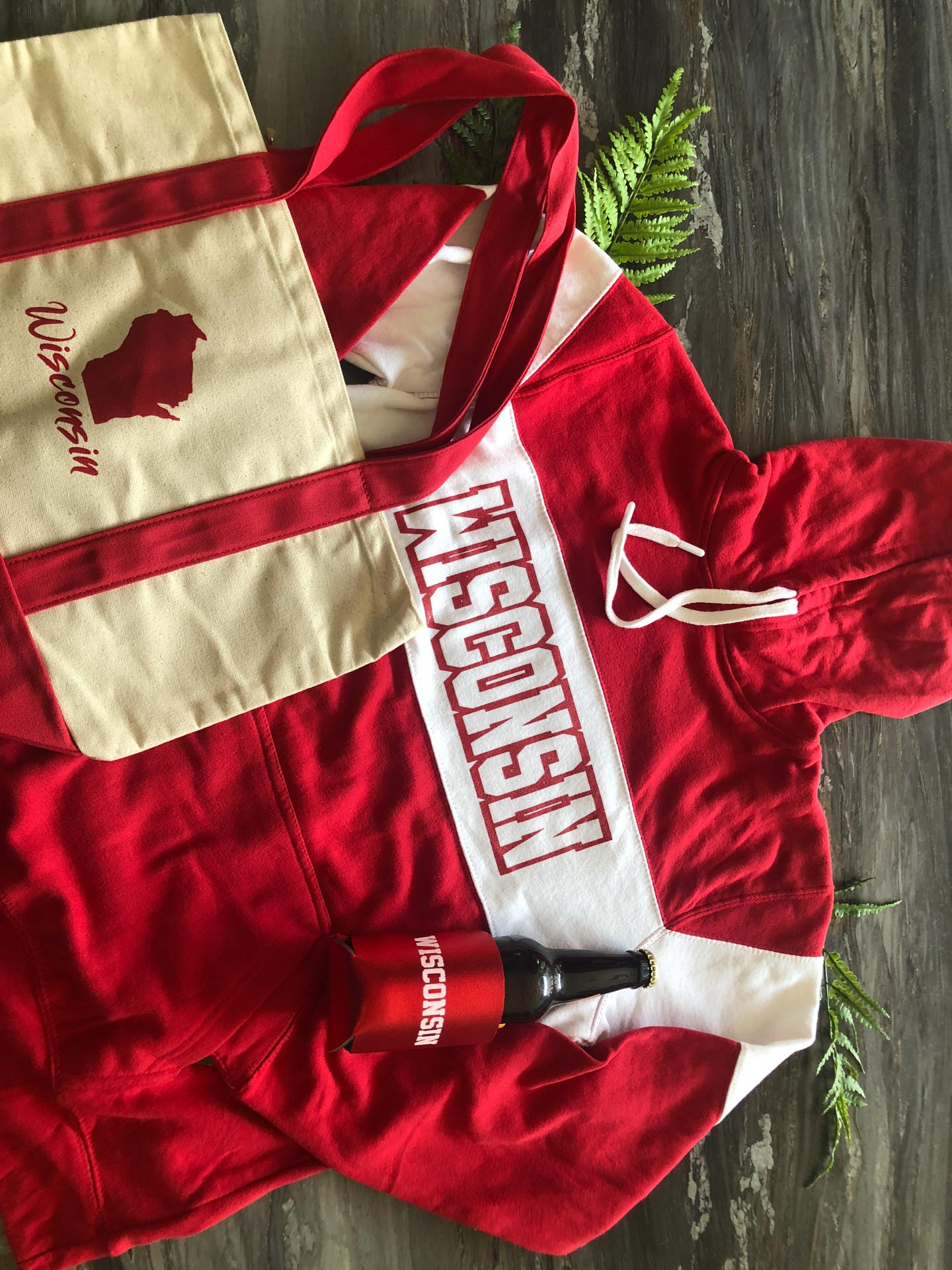 A red and white Wisconsin hoodie, a canvas tote bag with "Wisconsin" text and state outline, and a bottle in a red Wisconsin koozie on a wood surface with green ferns.