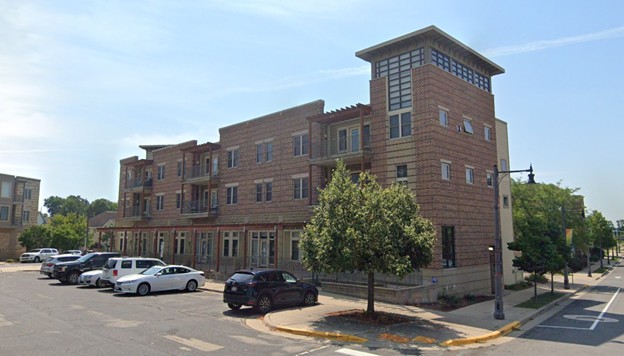 A three-story brick apartment building with balconies and a corner tower, adjacent to a parking lot with several parked cars and street trees.