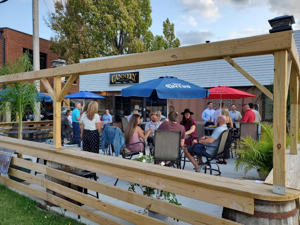 People sit and stand around tables under umbrellas on an outdoor patio at a restaurant or bar, with trees and a building in the background.