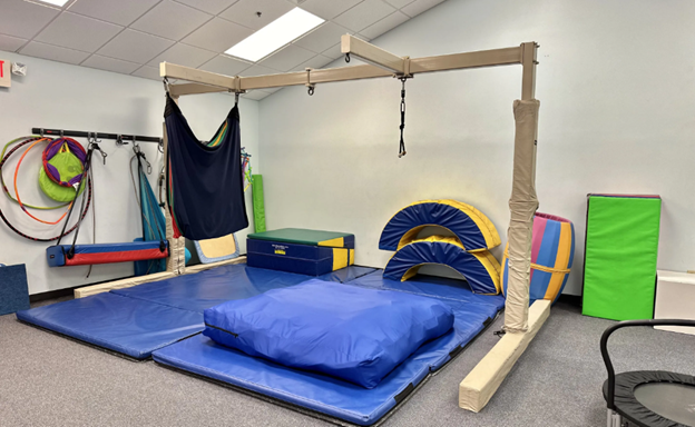 A therapy gym room with padded mats, a swing, climbing blocks, tunnels, balance equipment, and colorful foam shapes arranged against the walls.