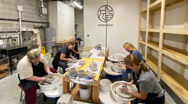 Four people seated at pottery wheels in a studio are shaping clay, with shelves and a sink visible in the background.