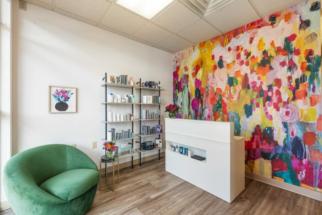 A modern reception area with a white desk, shelves of skincare products, a green chair, and a colorful abstract floral mural on the wall.