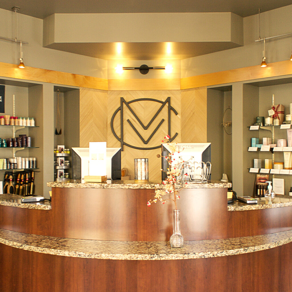 Reception desk at a modern salon or spa with two computer monitors, shelves displaying products, and a decorative wall logo behind the counter.