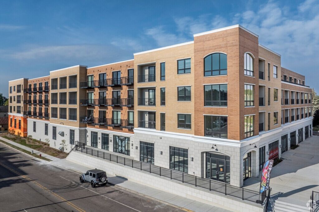 A modern three-story apartment building with brick and light-colored exterior, balconies, large windows, and ground-floor retail spaces along a city street.