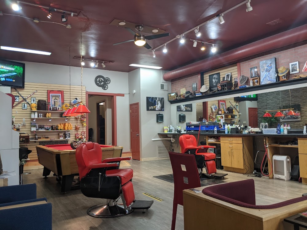 A barbershop interior with red chairs, a pool table, wall shelves holding various items, and large mirrors above wooden counters.