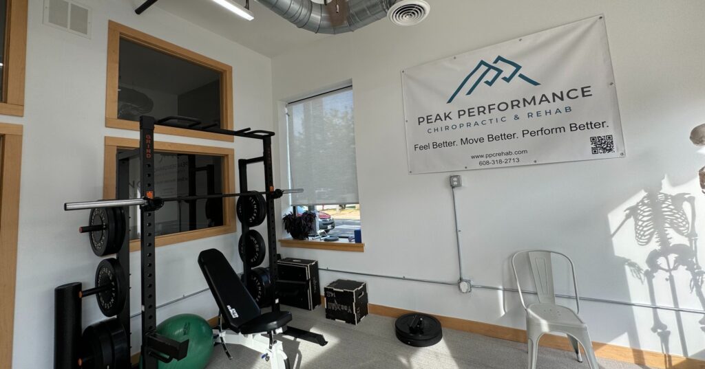 A gym corner in a rehab clinic with weightlifting equipment, a green exercise ball, a white chair, and a "Peak Performance Chiropractic & Rehab" banner on the wall.