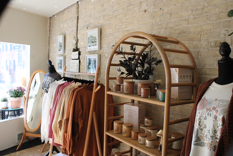 A boutique displays clothing on hangers, ceramic mugs and jars on shelves, framed art on a brick wall, and mannequins dressed in casual outfits.