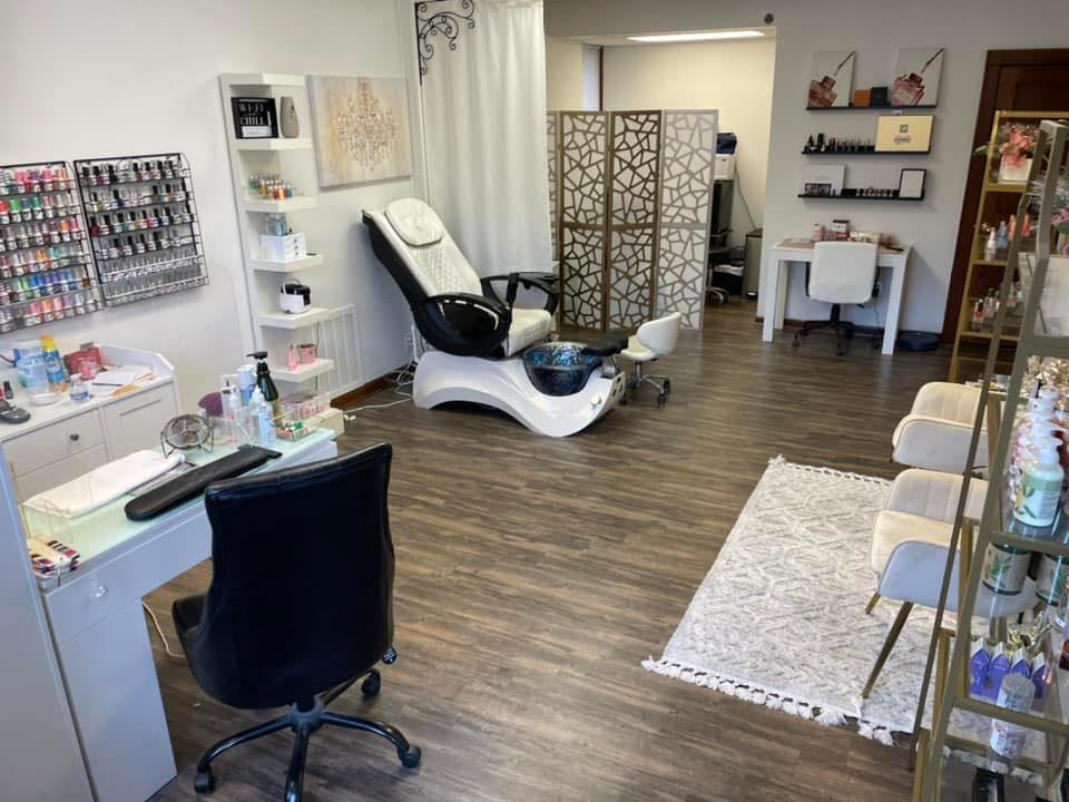 Nail salon interior with a pedicure chair, manicure stations, nail polish displays, and decorative partitions on wood flooring.