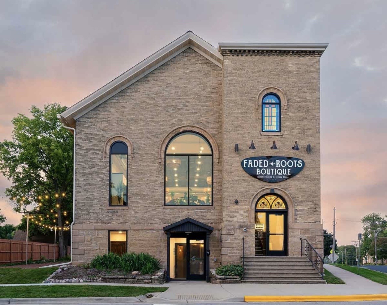 A two-story brick building with arched windows houses Faded Roots Boutique, as indicated by a sign near the entrance; exterior lights and landscaping are visible.