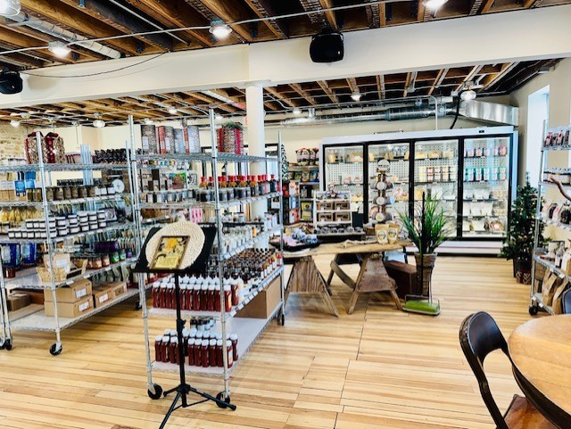 A brightly lit store interior with shelves stocked with various food items, refrigerated goods along the back wall, and a table display in the center on a wooden floor.