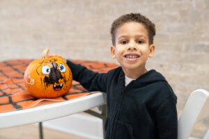 Young boy in a black hoodie smiles next to a small pumpkin decorated with marker drawings on a table covered with an orange and black cloth.