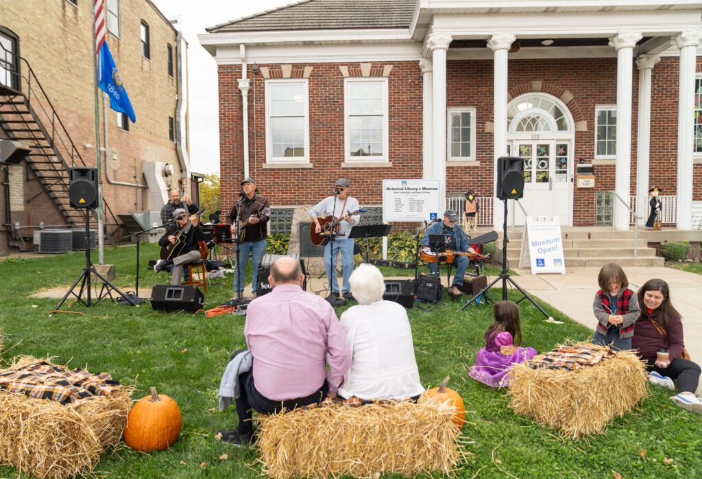 A band performs on a lawn in front of a brick building as people sit on hay bales and watch; pumpkins decorate the grass.