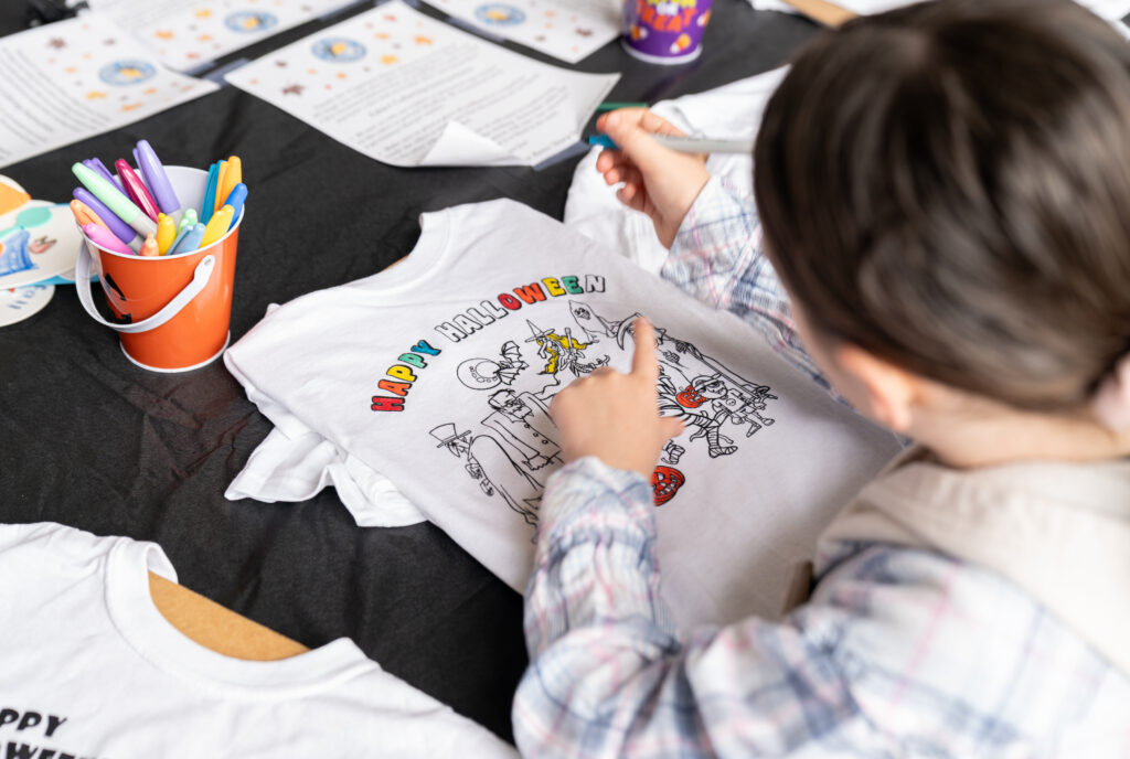 A child colors a “Happy Halloween” design on a white T-shirt using markers at a table with art supplies and printed instructions.