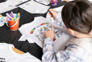 A child colors a “Happy Halloween” design on a white T-shirt using markers at a table with art supplies and printed instructions.