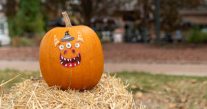 A pumpkin with cartoon monster face stickers sits on a straw bale outdoors, with a blurred background of trees and a building.