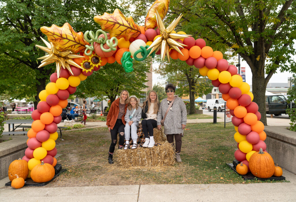 Four people pose on a hay bale under a colorful balloon arch decorated with pumpkins and autumn leaves at an outdoor event.