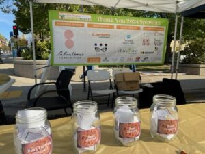 Four labeled jars filled with paper slips sit on a table under a tent, with a sponsor banner for the 2024 Farming Market displayed in the background.