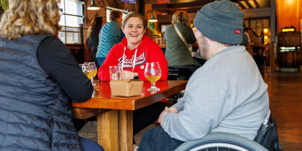 Three people sit around a wooden table in a casual indoor setting, talking and drinking beverages; one person uses a wheelchair.