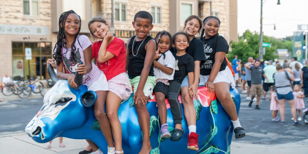 Seven children sit together on a painted blue cow sculpture outdoors, smiling at the camera with a crowd and buildings in the background.