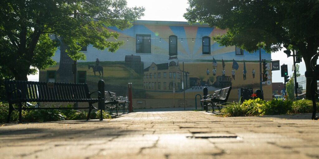 A mural depicting historical scenes is painted on the side of a building, with benches and trees in the foreground along a brick walkway.
