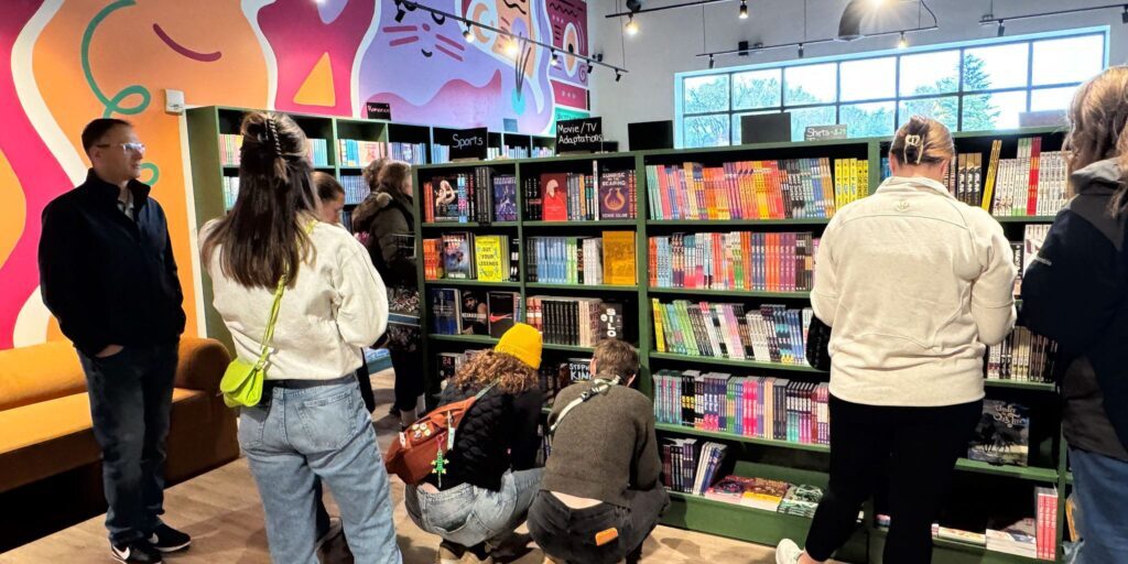 People browse colorful bookshelves in a brightly lit bookstore with vibrant, mural-covered walls. Some are standing, while others crouch to look at lower shelves.